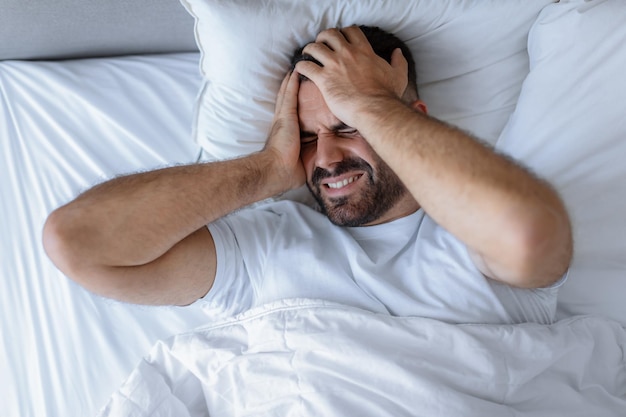 Hombre insomne acostado en la cama tocando la cabeza dolorida en casa.