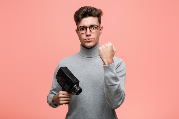 Hombre intelectual joven que sostiene una cámara de película que muestra el puño a la cámara, expresión facial agresiva.