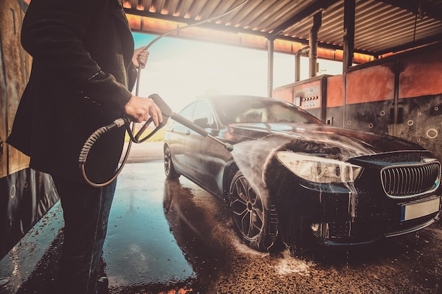 El hombre inteligente de moda en jeans y blaser está lavando su propio auto en la estación de lavado de autos.