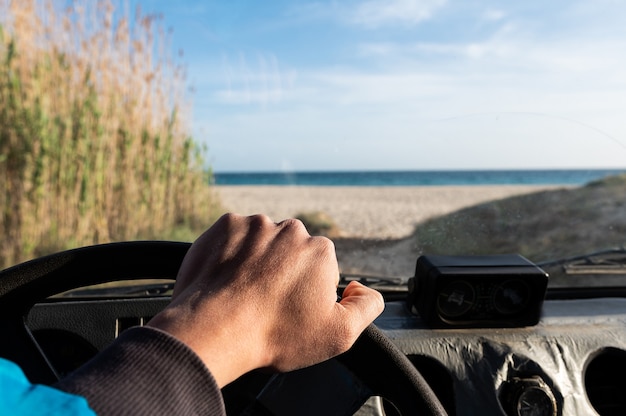 Hombre irreconocible con la mano en el volante mirando fuera de la ventana del coche con vistas a la hermosa playa de arena.