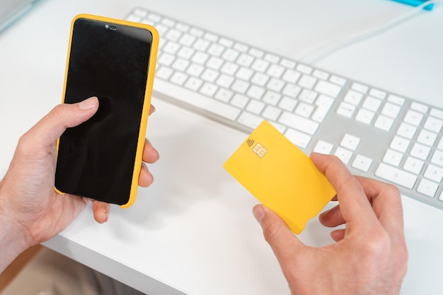 Hombre irreconocible pagando en línea con tarjeta de crédito y teléfono celular frente a una computadora