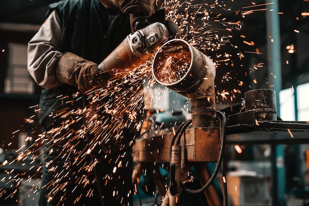 Hombre irreconocible en traje de protección y guantes de corte de tubería con amoladora. Interior del taller.