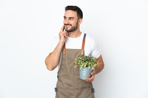 Hombre jardinero sosteniendo una planta aislada en la pared blanca pensando en una idea mientras mira hacia arriba