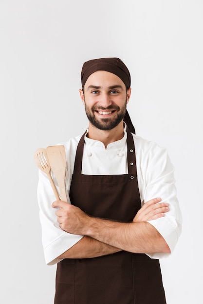 Hombre jefe guapo en uniforme de cocinero sonriendo mientras sostiene electrodomésticos de cocina de madera aislado sobre la pared blanca