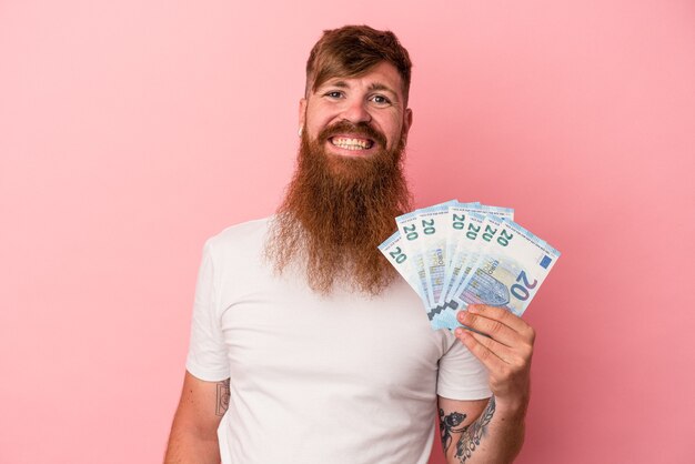 Hombre de jengibre caucásico joven con barba larga con billetes aislados sobre fondo rosa feliz, sonriente y alegre.