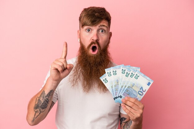 Hombre de jengibre caucásico joven con barba larga con billetes aislados sobre fondo rosa con una idea, concepto de inspiración.