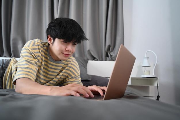 Hombre joven acostado en la cama y navegar por internet con ordenador portátil.