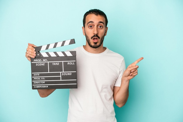 Hombre joven actor caucásico sosteniendo claqueta aislado sobre fondo azul apuntando hacia un lado