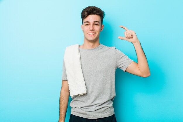 Foto hombre joven adolescente fitness sosteniendo algo poco con los dedos índices, sonriente y confiado