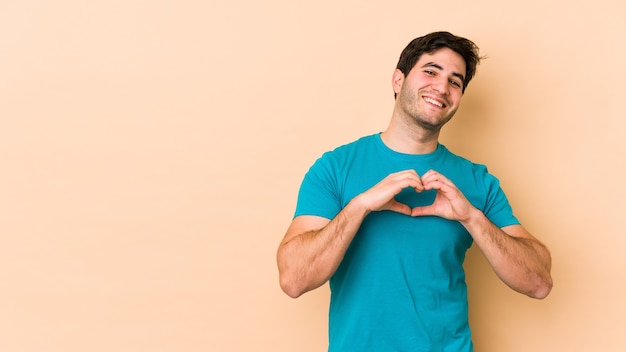 Hombre joven aislado en beige sonriendo y mostrando una forma de corazón con las manos.