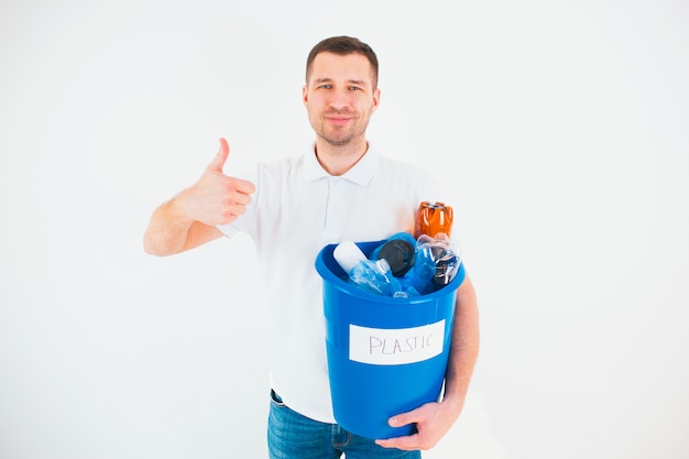 Hombre joven aislado sobre la pared blanca. Chico caucásico levanta el pulgar grande y sonríe. Cubo azul con botellas de plástico en el interior. Bueno para el proceso de reciclaje. Sin desperdicio de estilo de vida y medio ambiente.