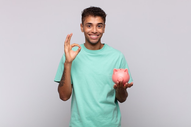 Hombre joven con una alcancía que se siente feliz, relajado y satisfecho, mostrando aprobación con gesto bien, sonriendo