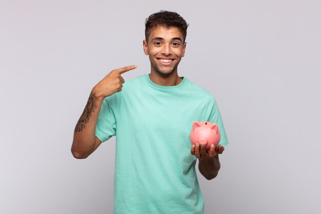 Hombre joven con una alcancía sonriendo con confianza apuntando a su propia sonrisa amplia, actitud positiva, relajada y satisfecha