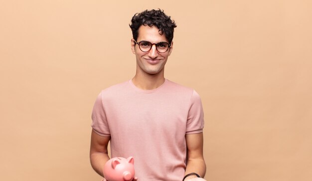 Hombre joven con una alcancía sonriendo felizmente con una mirada amistosa, segura y positiva, ofreciendo y mostrando un objeto o concepto