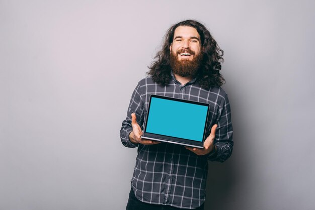 Hombre joven alegre barbudo hipster que muestra la pantalla del portátil en blanco sobre fondo gris