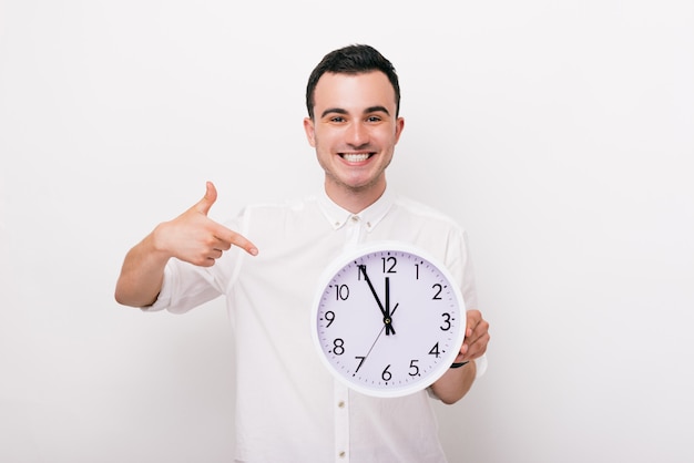 El hombre joven alegre está sonriendo y está mostrando un reloj blanco en un fondo blanco.