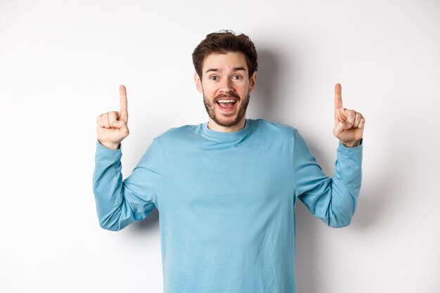 Hombre joven alegre que muestra publicidad con una sonrisa feliz, apuntando con el dedo hacia la pancarta del logotipo impresionante, de pie sobre fondo blanco.