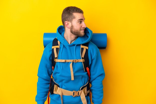 Hombre joven alpinista caucásico con una mochila grande aislada en amarillo mirando hacia el lado