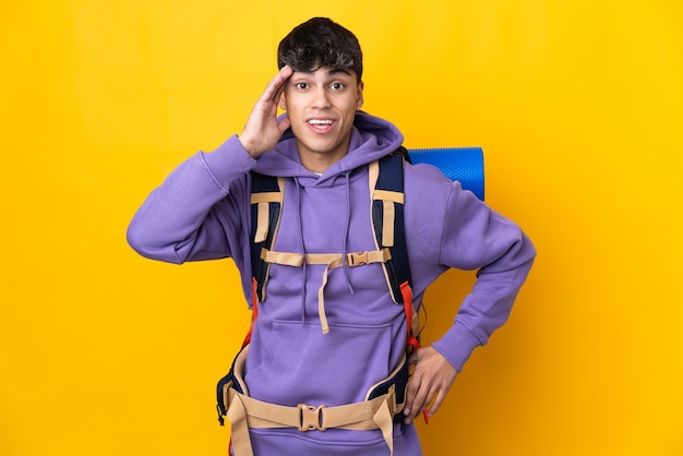 Hombre joven alpinista con una gran mochila sobre pared amarilla aislada con sorpresa y expresión facial conmocionada