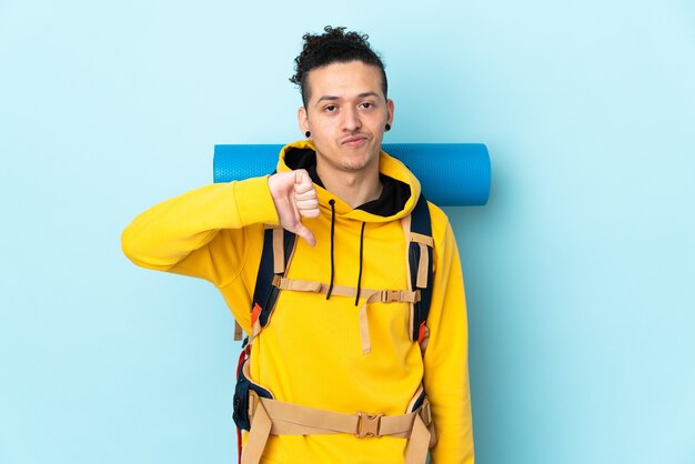 Hombre joven alpinista con una mochila grande sobre azul aislado mostrando el pulgar hacia abajo signo