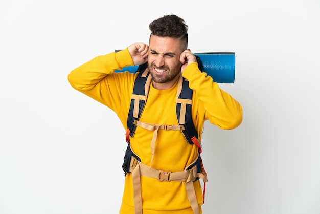 Hombre joven alpinista con una mochila grande sobre fondo aislado frustrado y cubriendo las orejas