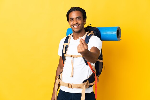 Hombre joven alpinista con trenzas con una mochila grande aislada en la pared amarilla haciendo gesto de dinero