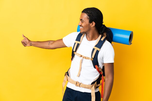 Hombre joven alpinista con trenzas con una mochila grande en amarillo dando un gesto de pulgar hacia arriba