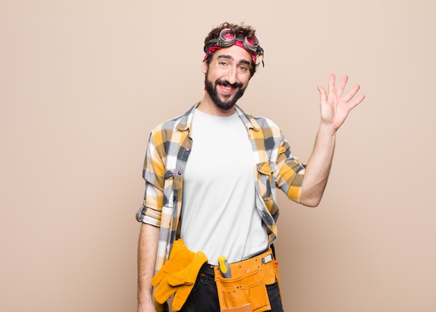 Hombre joven ama de llaves sonriendo feliz y alegremente, saludando con la mano, dándote la bienvenida y saludándote, o diciéndote adiós contra la pared plana