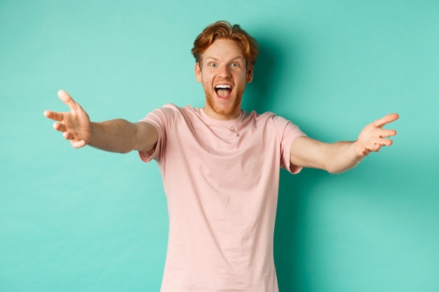 Hombre joven amable y feliz con cabello pelirrojo, estirar las manos en cálida bienvenida, alcanzar un abrazo y sonreír con alegría, de pie en camiseta sobre fondo de menta.