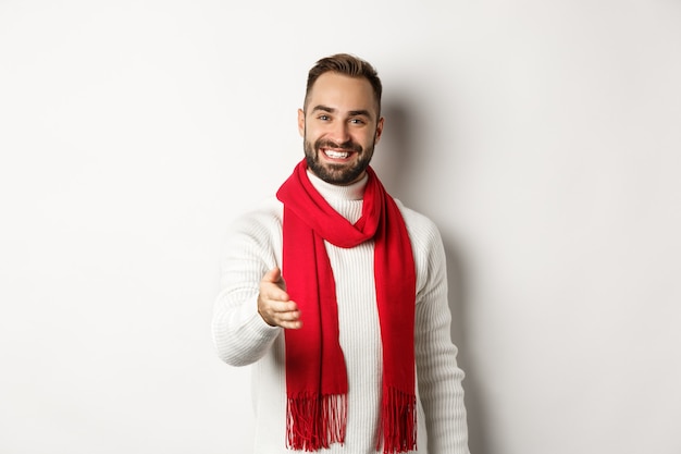 Hombre joven amable saludo persona con apretón de manos, sonriendo y deseando feliz navidad, vistiendo bufanda roja y suéter de invierno, fondo blanco.