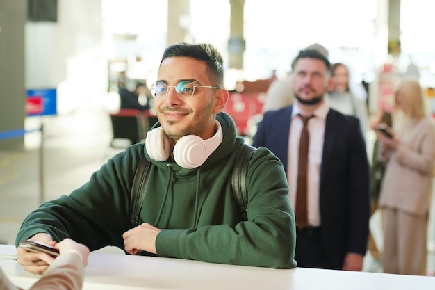 Hombre joven en anteojos de pie en la recepción y el check-in antes del vuelo con personas de pie detrás de él en el fondo