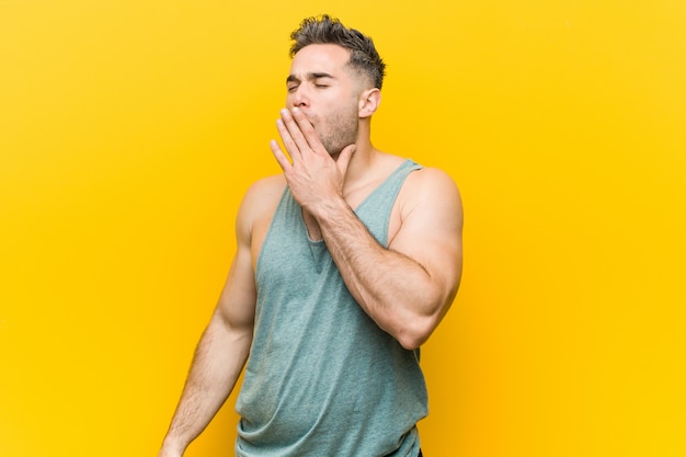 Hombre joven de la aptitud contra una pared amarilla que bosteza mostrando un gesto cansado que cubre la boca con la mano.