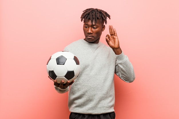 Hombre joven de la aptitud del negro que sostiene un balón de fútbol alegre y confiado que muestra gesto aceptable.