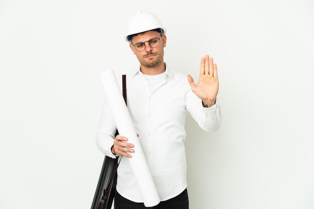 Hombre joven arquitecto con casco y sosteniendo planos aislados en blanco haciendo gesto de parada y decepcionado