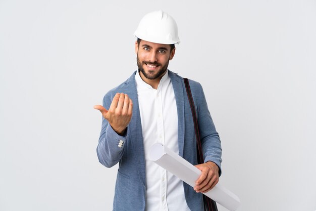 Hombre joven arquitecto con casco y sosteniendo planos aislados en blanco invitando a venir con la mano. Feliz de que hayas venido