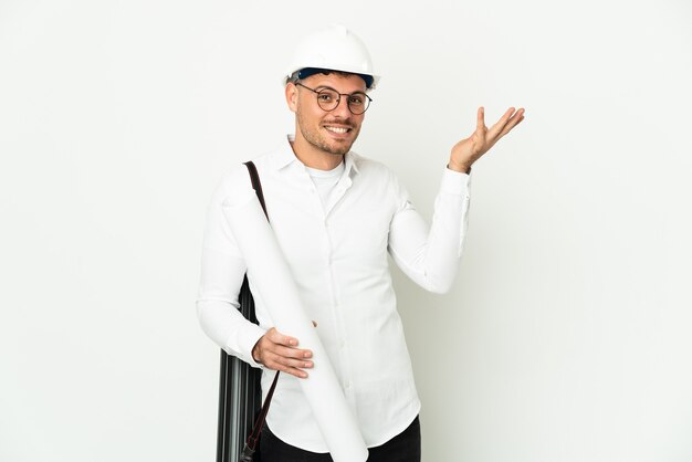 Hombre joven arquitecto con casco y sosteniendo planos aislados en la pared blanca extendiendo las manos hacia un lado para invitar a venir