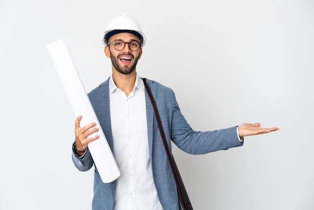 Hombre joven arquitecto con casco y sosteniendo planos aislados en la pared blanca extendiendo las manos hacia un lado para invitar a venir