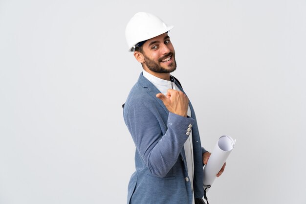 Hombre joven arquitecto con casco y sosteniendo planos aislados sobre fondo blanco apuntando hacia el lado para presentar un producto