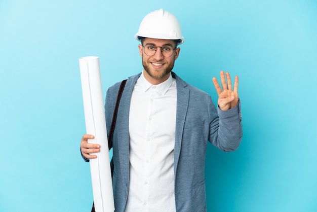 Hombre joven arquitecto con casco y sosteniendo planos sobre fondo aislado feliz y contando cuatro con los dedos