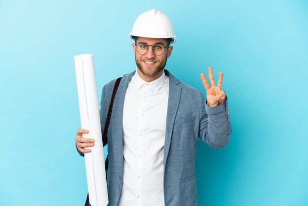 Hombre joven arquitecto con casco y sosteniendo planos sobre pared aislada feliz y contando tres con los dedos