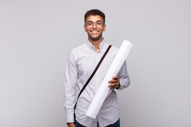 Hombre joven arquitecto sonriendo felizmente con una mano en la cadera y actitud confiada, positiva, orgullosa y amistosa