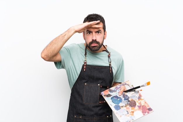 Hombre joven artista sosteniendo una paleta sobre pared aislada mirando lejos con la mano para mirar algo