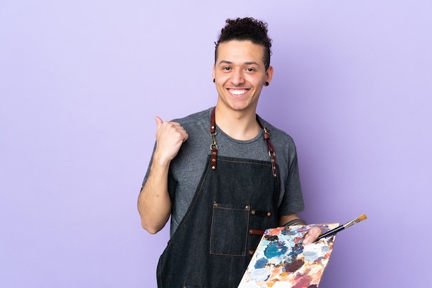Hombre joven artista sosteniendo una paleta sobre pared púrpura aislada apuntando hacia el lado para presentar un producto