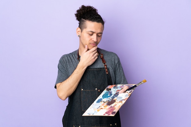Hombre joven artista sosteniendo una paleta sobre pared púrpura aislada con expresión triste