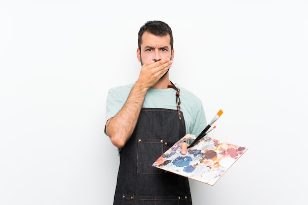 Hombre joven artista sosteniendo una paleta sobre paredes aisladas que cubren la boca con las manos
