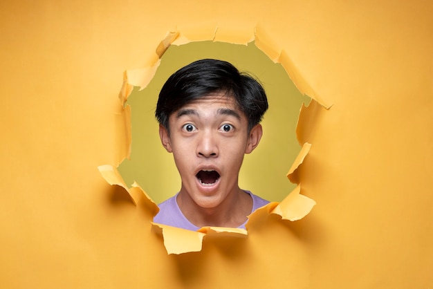 Hombre joven asiático con expresión de asombro y asombro hombre adolescente, posa a través del agujero de papel amarillo rasgado, vistiendo una camiseta púrpura, asustado y conmocionado con expresión de sorpresa, miedo y cara emocionada