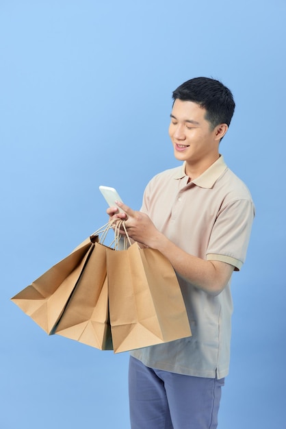 Hombre joven asiático sosteniendo bolsas de la compra y usando teléfono celular, retrato de cuerpo entero aislado.