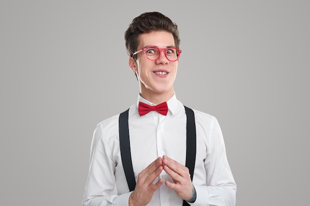 Hombre joven astuto con gafas rojas mirando a la cámara con una sonrisa engañosa y haciendo un gesto de campanario mientras planea contra el fondo gris