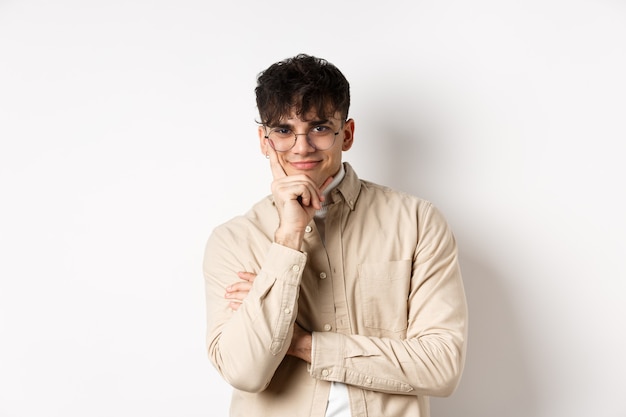 Hombre joven astuto que ve pasar algo interesante, con la cara apoyada en el brazo y una sonrisa tortuosa, intrigada, de pie sobre una pared blanca.