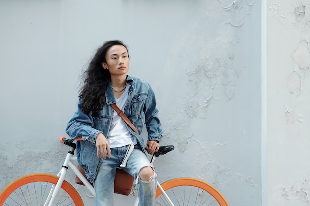 Hombre joven atractivo elegante en jeans y chaqueta de mezclilla apoyado en bicicleta y mirando a otro lado